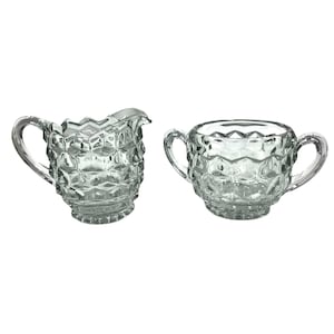 May include: A clear glass creamer and sugar bowl set. Both pieces have a geometric pattern and curved handles. The creamer has a spout. The sugar bowl has two handles. The set is likely vintage and suitable for serving.