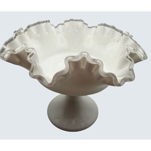 Puede incluir: Un cuenco de pedestal de vidrio blanco con un borde con volantes. El cuenco tiene un diseño ancho y poco profundo y se asienta sobre una base redonda. El borde tiene un borde transparente y ligeramente texturizado.