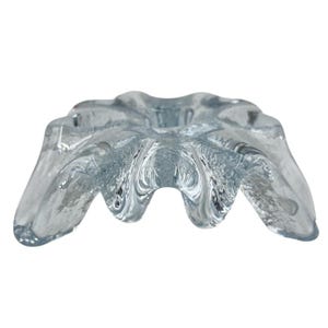 Puede incluir: Un cuenco de vidrio transparente de color azul claro con una forma orgánica única. El cuenco tiene una superficie texturizada y una depresión central, con bordes ondulados que crean una apariencia de estrella. El cuenco probablemente se utiliza con fines decorativos.