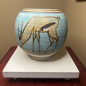 May include: A white ceramic mug with a black handle and the text "But First, Coffee" in a bold, black font. The mug is set against a blurred background, suggesting a cozy setting.