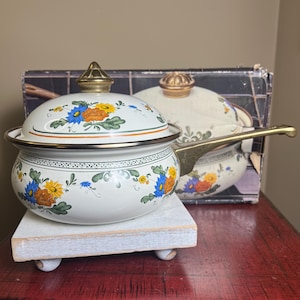 May include: A white enameled metal saucepan with a matching lid, featuring a floral pattern of blue, orange, and yellow flowers with green leaves. The pot has a brass-toned metal handle and a matching brass-toned knob on the lid.
