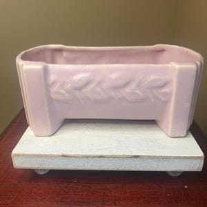 May include: A rectangular, light pink ceramic planter with a leaf design. The planter sits on a white, wooden stand. The planter has a rectangular shape with a decorative leaf pattern in the center.