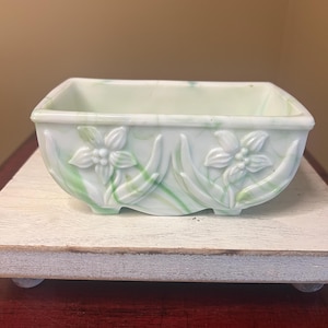 May include: A rectangular, light green and white decorative dish with a floral design. The dish has a square shape and is decorated with embossed flowers and leaves. The color is a marbled effect of white and light green.
