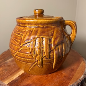 May include: A round, amber-colored ceramic jar with a matching lid and a single side handle. The exterior features a raised, textured relief pattern of elongated leaves or pods wrapping around the body, finished in a glossy, mottled brown glaze.