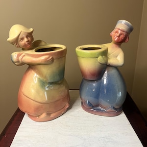 May include: Two vintage ceramic figurines of figures holding flower pots. The figurines are painted in pastel colors, with one in shades of yellow, orange, and blue, and the other in blue, pink, and yellow. The figures have detailed faces and are posed as if they are embracing the pots.
