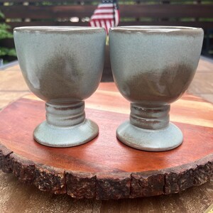 May include: Two ceramic goblets with a light blue-grey glaze and brown mottled accents. Each goblet features a wide bowl, a short stem with two horizontal ridges, and a circular base. A wooden ruler is visible at the bottom right corner.