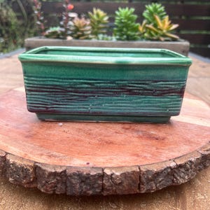 May include: A rectangular ceramic planter with a glossy green glaze and dark brown horizontal textured bands. The vessel features a wide opening, a flat rim, and a slightly tapered base, showcasing a reactive finish with subtle color variations.