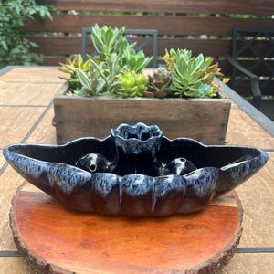 May include: A dark blue ceramic planter with a unique shape, featuring a glossy finish and a light blue drip glaze. The planter sits on a wooden slice, with a wooden box of succulents in the background.