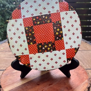 May include: A circular metal tin lid featuring a patchwork quilt pattern. The design consists of alternating squares in cream with small red floral motifs, bright red with white polka dots, and dark brown with yellow and red floral patterns.