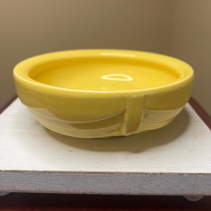 May include: A small, round, yellow ceramic bowl with a smooth, glossy finish. The bowl has a shallow interior and a slightly raised rim. The exterior features a subtle, textured design. The bowl is sitting on a white and gray wooden surface.