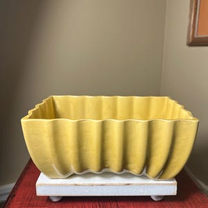 May include: A yellow ceramic planter with a ribbed design. The rectangular planter sits on a white wooden base with small, round feet. The planter is a warm, mustard yellow color.