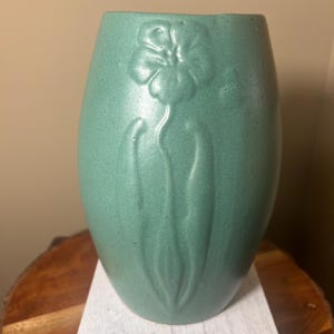 May include: A teal-colored vase with a textured surface and a raised floral design. The vase has an oval shape and is displayed on a white wooden block. The floral design is centered on the front of the vase.