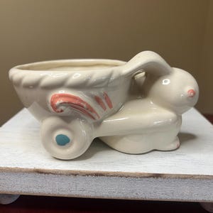 May include: Cream-colored ceramic planter shaped like a bunny pulling a cart. The cart has a scalloped rim and pink and red accents. The bunny has a pink nose and blue wheel.