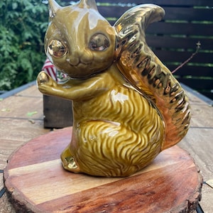May include: A ceramic squirrel figurine featuring a glossy olive-green finish on the body and a reflective metallic gold glaze on the large, curved tail and eyes. The squirrel is depicted in a seated position with one paw raised forward.