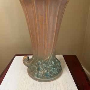 May include: A tall, fluted vase with a flared top. The vase is primarily a muted peach color with vertical lines and a teal-colored base adorned with a cluster of stylized fruit. The vase has a curved, scroll-like detail on one side.