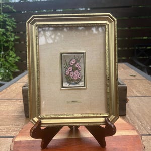 May include: A rectangular gold-toned frame with a distressed finish containing a centered, small, vertical metallic plaque featuring a relief of pink flowers in a vase. The plaque includes the text L. Marque and is set against a beige fabric mat.