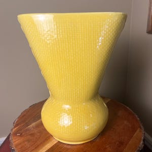 May include: A bright yellow ceramic vase with a flared top and a rounded base. The exterior surface features a distinct, uniform grid-like texture that covers the entire body of the vessel, creating a subtle pattern across the glossy finish.