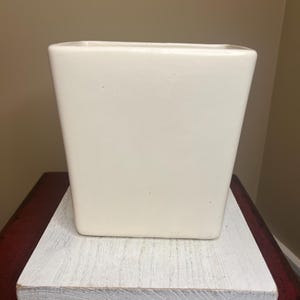 May include: A square, off-white ceramic planter sits on a white-washed wooden base. The planter has a slightly tapered design. The base rests on a dark red surface, creating a contrast in colors and textures.