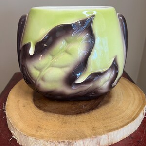 May include: A ceramic planter with a glossy finish, featuring a design of stylized leaves. The planter has a light green interior and is adorned with dark purple leaves. It sits on a round, wooden slice.