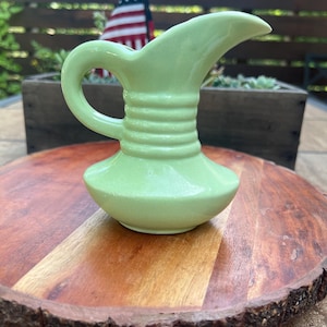 May include: A light green ceramic pitcher featuring a glossy finish, a curved handle, a wide pouring spout, and three horizontal raised ridges encircling the neck of the vessel. The pitcher has a rounded base that tapers upward to the spout.