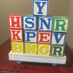May include: Wooden alphabet blocks in various colors, including red, blue, green, and yellow. Each block features a letter of the alphabet. The blocks are stacked on a small, white, rectangular base with small, round feet.
