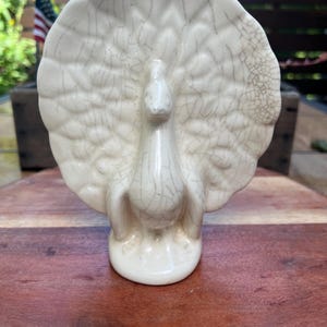 May include: A cream-colored ceramic peacock figurine featuring a prominent fanned tail and a distinct crackle glaze finish. The bird stands on a rounded base, with the textured, webbed pattern of the glaze visible across the entire surface.