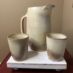 May include: A beige ceramic pitcher with a handle and two matching cups sit on a white wooden tray. The pitcher is tall and cylindrical, and the cups are small and rounded. The set is displayed on a red surface.