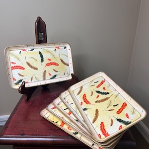 May include: Rectangular serving trays with a feather design. The trays have a cream-colored background with scattered red, brown, and black feathers. The edges are gold-colored. One tray is displayed on a wooden stand, while the others are stacked.