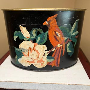 May include: A black metal container with a gold rim, featuring a hand-painted design of a red cardinal bird perched on a branch with a large white and orange flower and green leaves. The container is sitting on a white wooden surface.