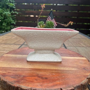 May include: A ceramic planter featuring a textured, off-white exterior finish and a smooth, light pink interior. The vessel has a wide, rectangular opening that tapers toward a central pedestal base, creating a distinct, elongated silhouette.