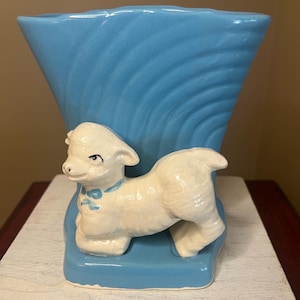 May include: A vintage ceramic vase in a light blue hue, featuring a lamb figurine. The vase has a textured, wave-like design. The lamb is white with a blue bow, adding a charming, pastoral touch. A decorative piece.