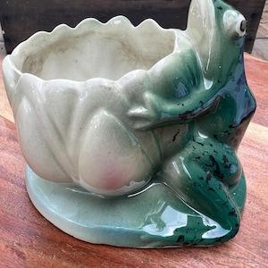 May include: A ceramic planter shaped like a lily pad with a green frog attached to the side. The bowl of the planter is off-white with a scalloped rim, while the frog is dark green with black speckles. The entire piece sits on a flat base.