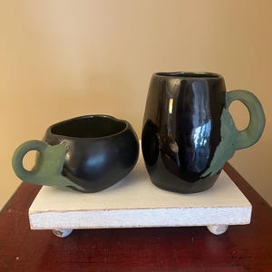 May include: Two black ceramic mugs with green handles. The mugs are shaped like eggplants. The mugs are on a white wooden tray with rounded edges.