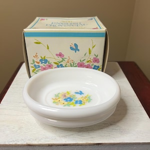 May include: A white, round, shallow dish with a floral design in the center, featuring blue, yellow, and pink flowers and a blue butterfly. The dish sits on a white wooden surface, with a box behind it that reads "ENGLISH PROVINCIAL".