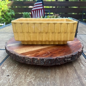 May include: A rectangular mustard yellow ceramic planter featuring a vertical ribbed texture along the exterior walls. The vessel has a stepped base design and a wide, open top, presenting a solid, uniform color throughout its glazed surface.