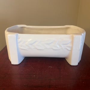 May include: A rectangular, white ceramic planter with a raised leaf design on the front. The planter has a slightly curved top and is supported by two short feet. The surface is smooth and glossy, suitable for indoor or outdoor use.