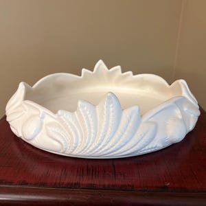 May include: A white ceramic bowl with a low profile and a scalloped rim. The exterior features a relief pattern of stylized leaves and rounded fruit shapes arranged in a repeating design around the entire circumference of the circular vessel.