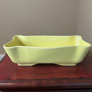 May include: A yellow ceramic planter with a rectangular shape and slightly flared edges. The planter has four small feet on the bottom.