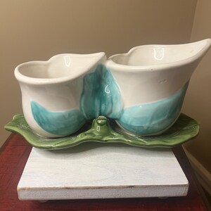 May include: A white ceramic serving dish with two compartments, each with a curved rim and teal accents. The dish sits on a green leaf-shaped base, perfect for serving dips or snacks. The dish is placed on a white wooden surface.