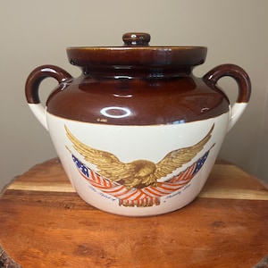 May include: Brown and white ceramic crock with a lid and handles. The crock features a gold eagle design with red, white, and blue accents. The text "The Spirit of Seventy Six" is printed below the eagle.
