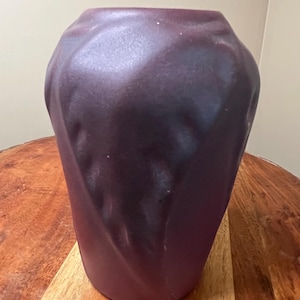 May include: A dark purple vase with a textured, geometric design. The vase has a wide base and tapers slightly towards the top. The surface has a matte finish. The vase is sitting on a wooden surface.