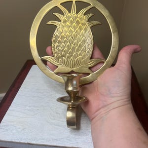 May include: A brass pineapple wall sconce. The pineapple is centered within a circular frame, with a candle holder below. The sconce is held by a hand, showcasing its size and design. The pineapple design is a popular decorative motif.