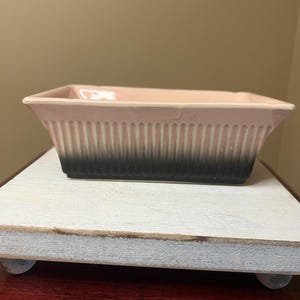 May include: A rectangular, pink ceramic baking dish with vertical ribbed detailing and a gradient from pink to black at the base. The dish is displayed on a white, wooden stand with small, round feet.