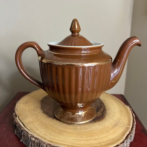 May include: A brown ceramic teapot with gold accents. The teapot has a rounded body with vertical lines and a lid with a knob on top. The teapot is sitting on a wooden base.