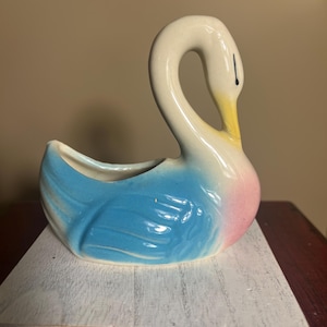 May include: A ceramic swan planter with a curved neck and open top. The swan is painted with a gradient of colors, including white, yellow, pink, and blue. The planter is sitting on a white wooden base.
