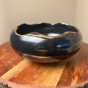May include: A round, black ceramic bowl with a wavy, uneven rim. The exterior surface features horizontal, brushed gold paint strokes that wrap around the body, contrasting against the matte black finish of the bowl's exterior and interior.