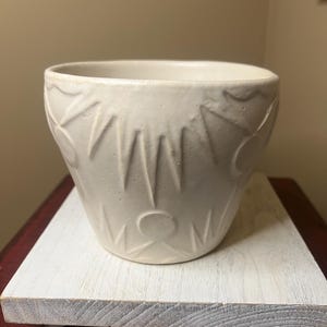 May include: Off-white ceramic planter with a textured, carved design. The planter has a wide opening and a slightly tapered shape. The carved pattern features vertical lines and circular shapes. The planter sits on a white wooden surface.
