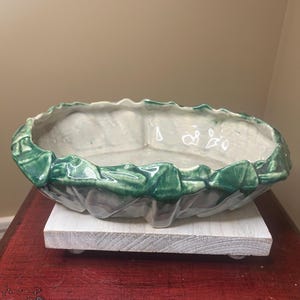 May include: An oval-shaped ceramic planter with a green, leaf-like trim. The interior is a light gray color. The planter sits on a white, rectangular wooden base. The base is on a dark red surface.