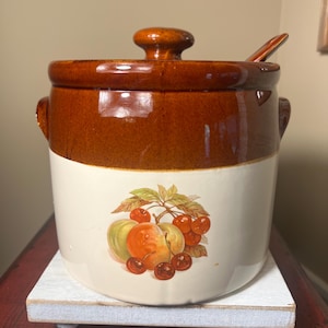 May include: A two-tone ceramic jar with a lid and a small spoon. The top is brown, and the bottom is white with a fruit illustration. The jar sits on a small, white wooden stand. The jar has two small handles.