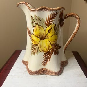 May include: Cream-colored ceramic pitcher with a large yellow flower design. The pitcher has a ruffled rim and base, with brown trim and a curved handle. The floral design includes green leaves and brown accents.